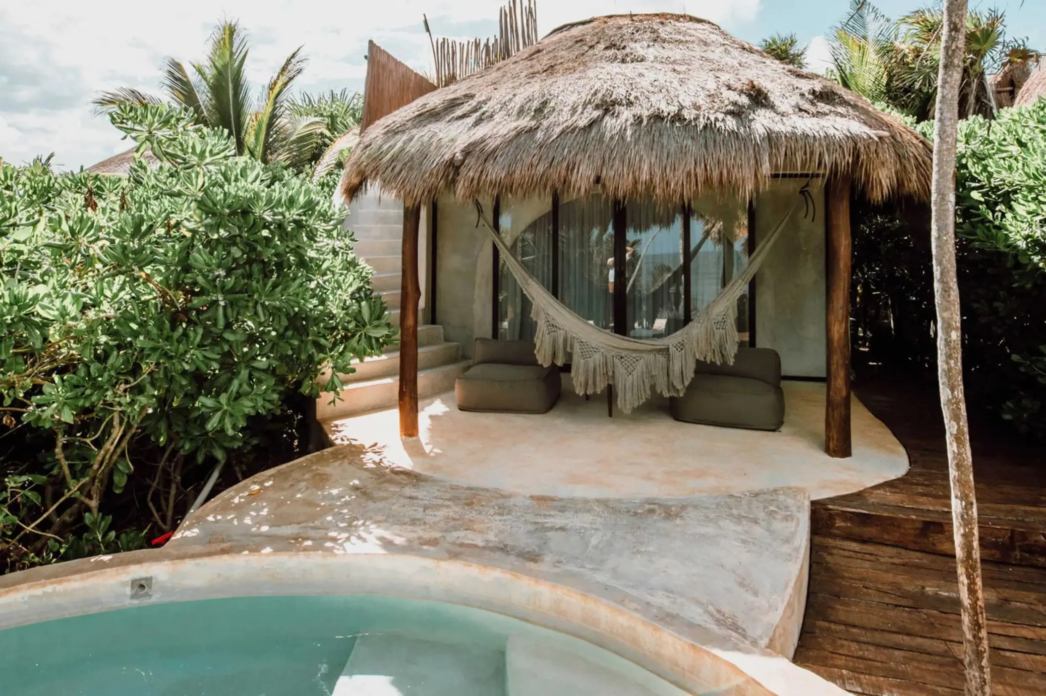 A charming casita with a thatched roof, hammock, small plunge pool, and lush greenery, offering an ocean view.