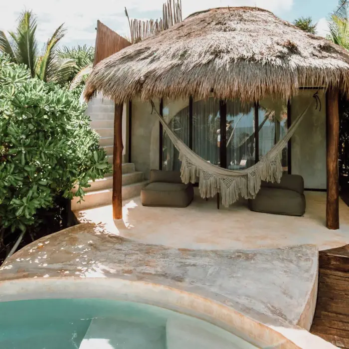 A charming casita with a thatched roof, hammock, small plunge pool, and lush greenery, offering an ocean view.
