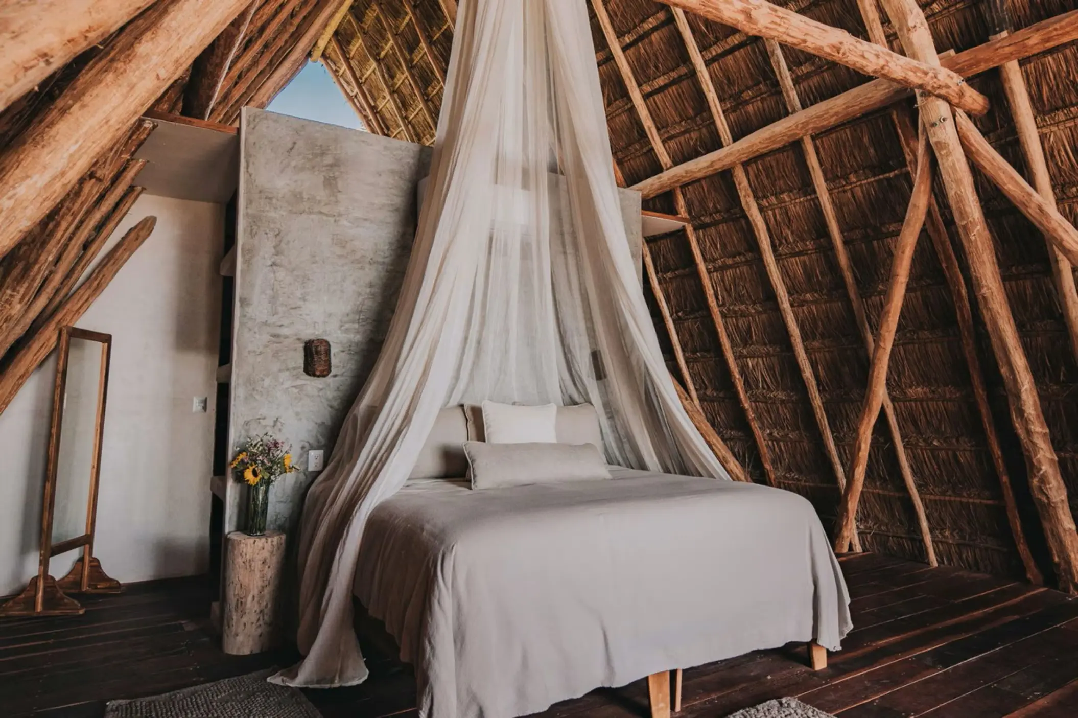 Interior of a loft with a high thatched roof, featuring a king bed draped in a white mosquito net and wooden floors.