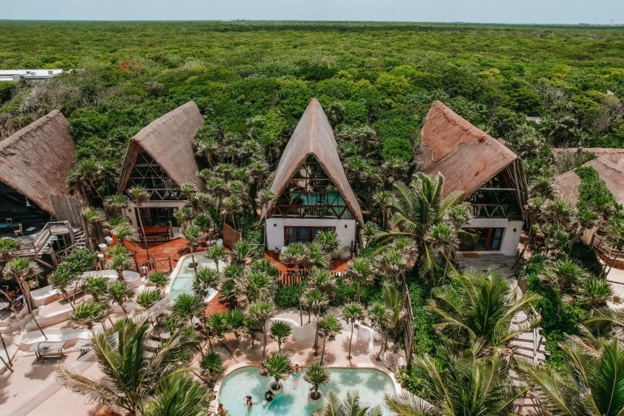 Aerial view of A-frame loft structures with thatched roofs, surrounded by palm trees and a central swimming pool area.