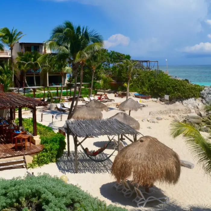 A picturesque beach scene at Piedra Escondida with palm trees, thatched umbrellas, lounge chairs, and clear turquoise ocean.