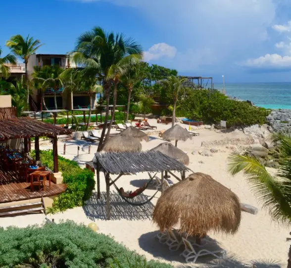 A picturesque beach scene at Piedra Escondida with palm trees, thatched umbrellas, lounge chairs, and clear turquoise ocean.