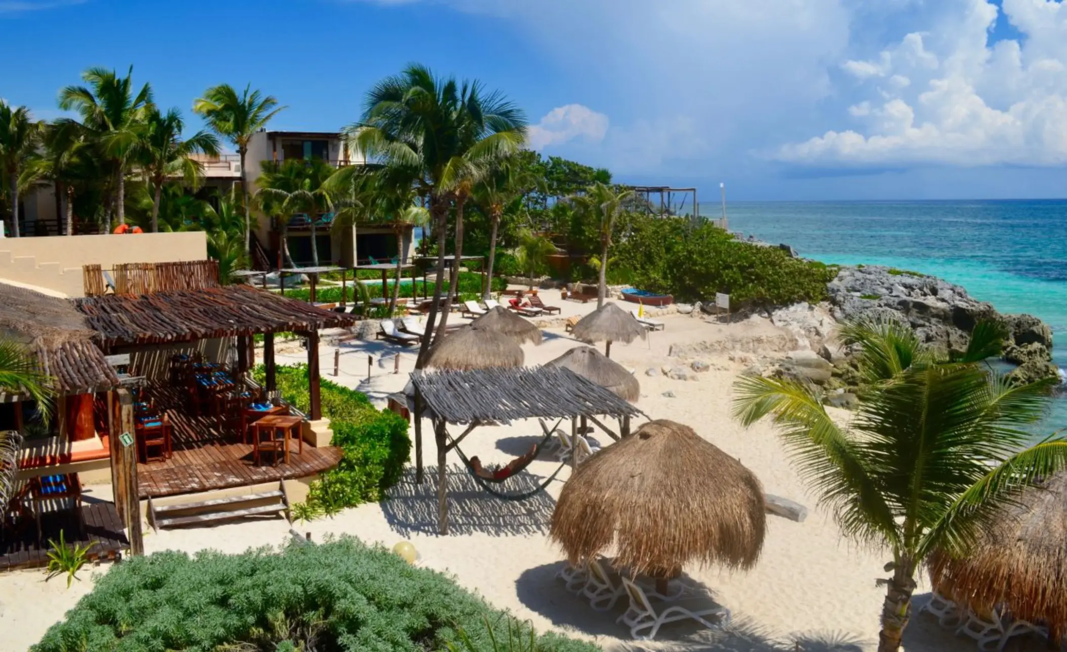 A picturesque beach scene at Piedra Escondida with palm trees, thatched umbrellas, lounge chairs, and clear turquoise ocean.