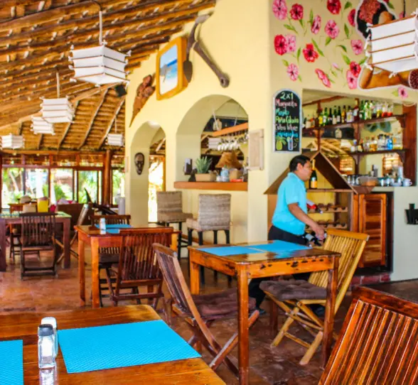 Interior of Restaurant Molcajete at Piedra Escondida with a thatched roof, wooden tables, a bar, and a server in a blue shirt.