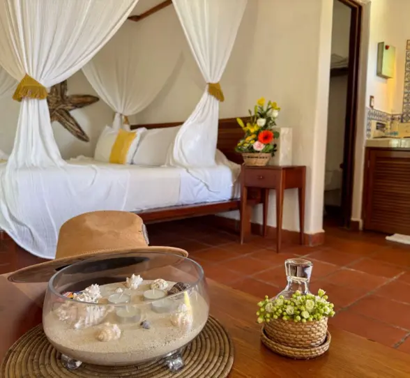 A cozy hotel room at Piedra Escondida with a four-poster bed draped in white, a decorative bowl, and a colorful sink.