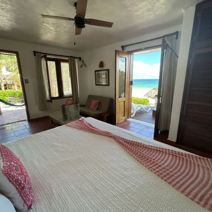 A bright hotel room at Piedra Escondida with a large bed, ceiling fan, and open doors leading to a balcony with an ocean view.