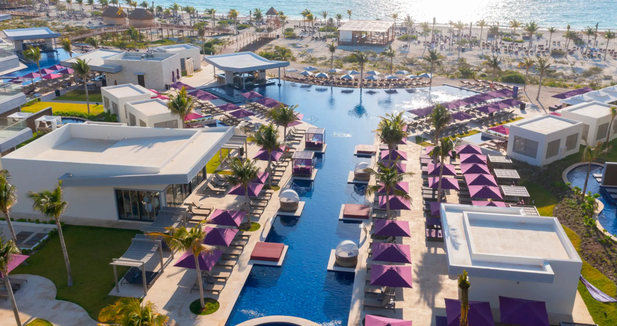 Aerial view of a beachfront resort with central infinity pool, cabanas, and purple umbrellas lining both sides.