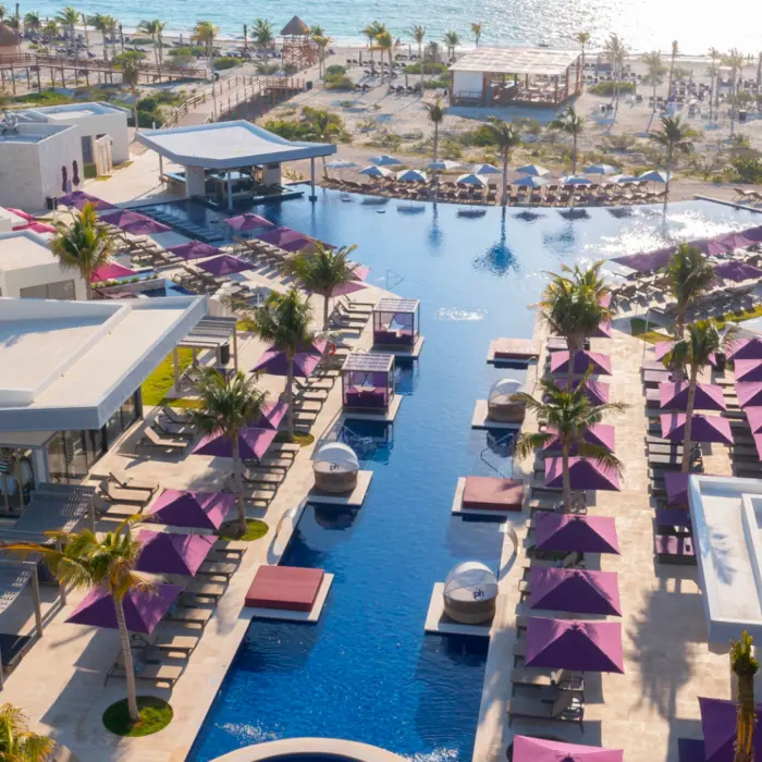 Aerial view of a beachfront resort with central infinity pool, cabanas, and purple umbrellas lining both sides.