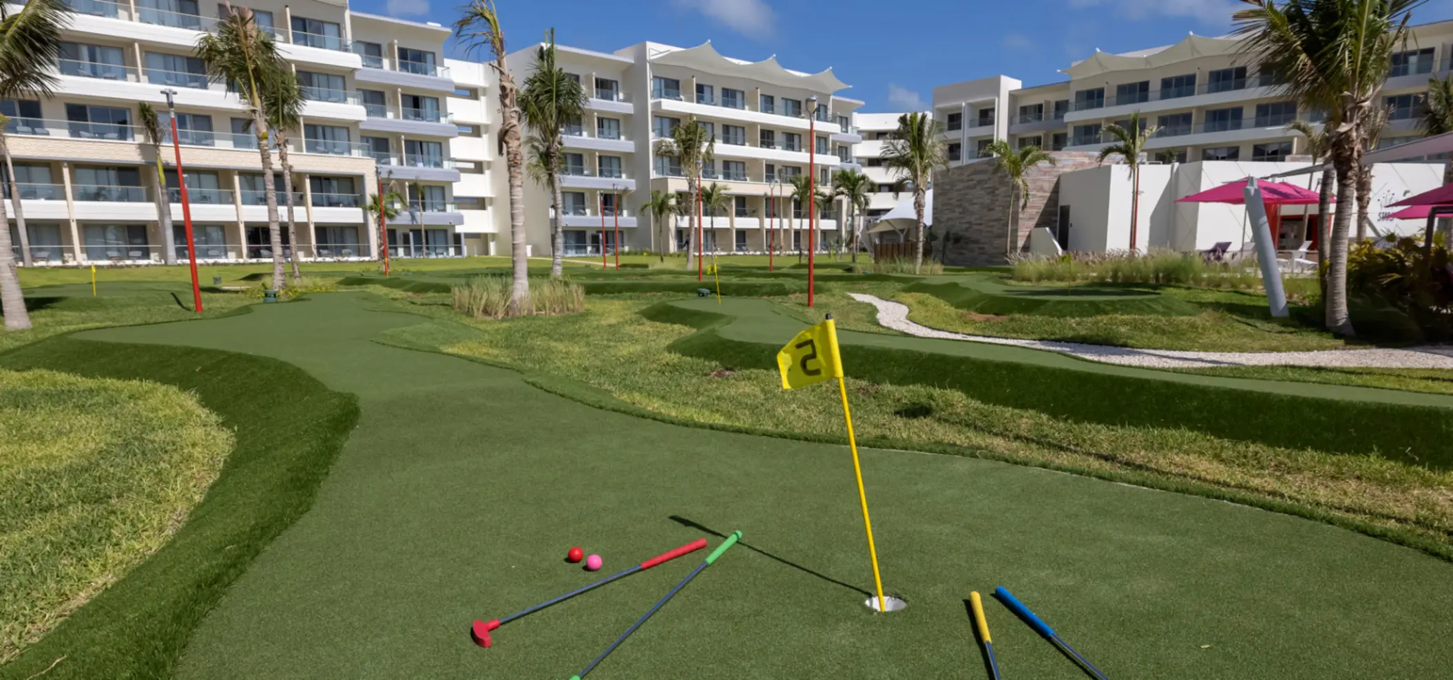Mini-golf course with colorful putters on green turf, surrounded by palm trees and resort buildings.