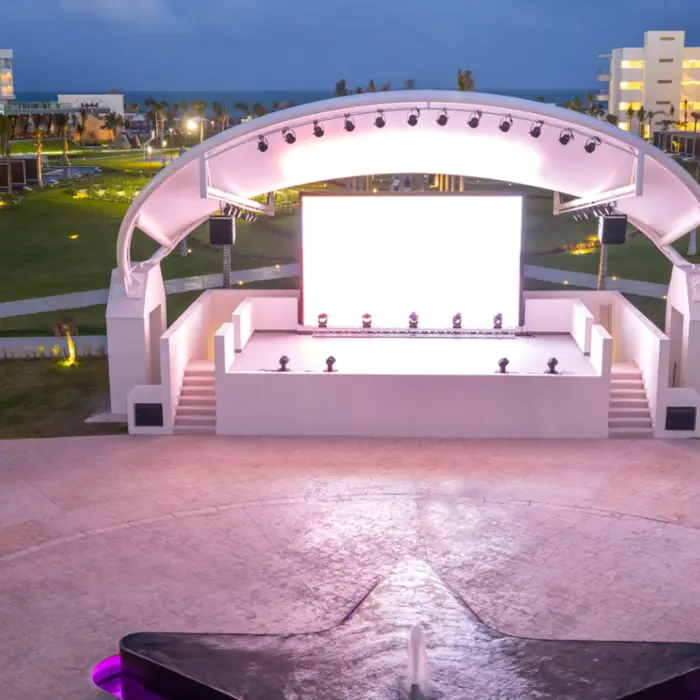 Open-air stage with glowing lights and large screen, set between modern resort buildings at twilight.