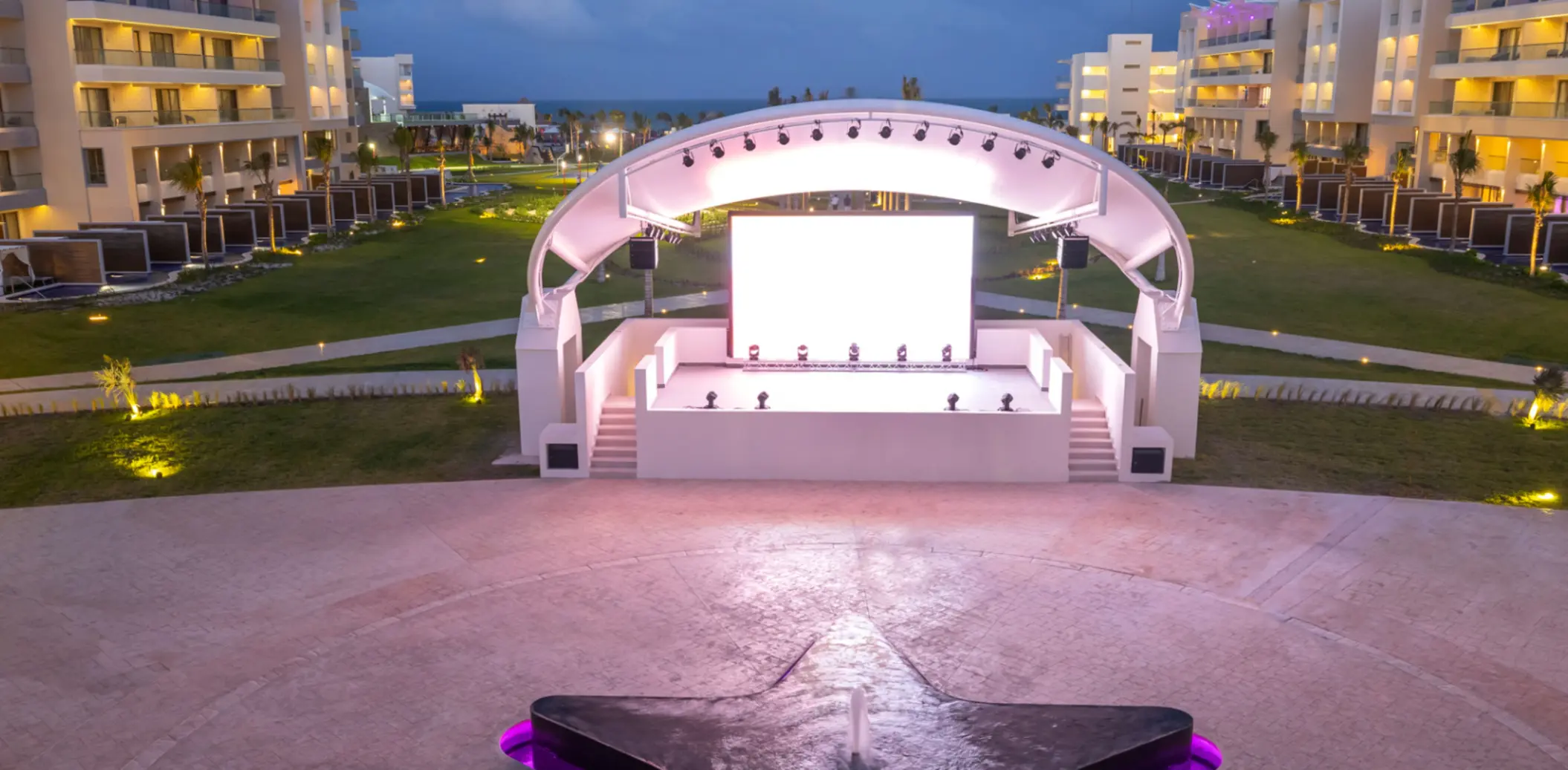 Open-air stage with glowing lights and large screen, set between modern resort buildings at twilight.