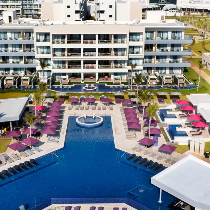 Symmetrical view of resort buildings and pool area with lounge chairs and cabanas under purple umbrellas.