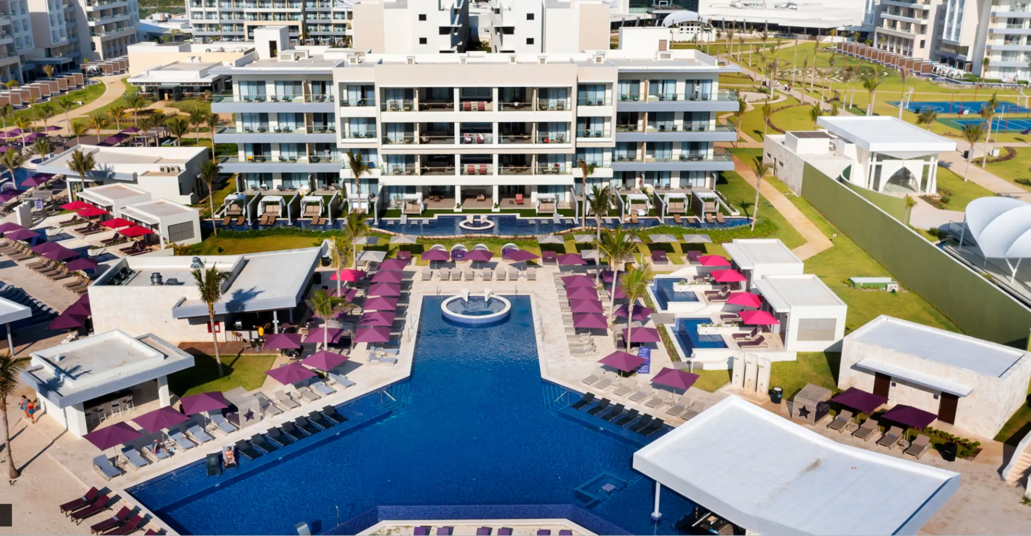 Symmetrical view of resort buildings and pool area with lounge chairs and cabanas under purple umbrellas.