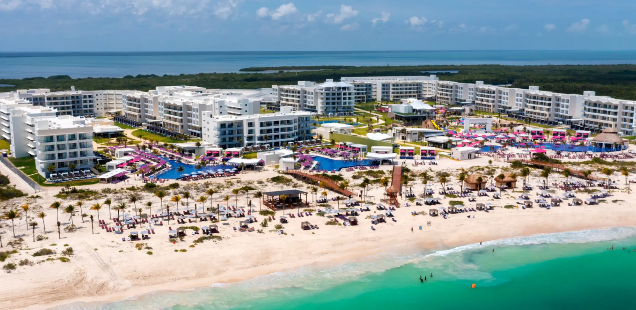Wide aerial shot of beachfront resort with multiple pools, white buildings, and turquoise sea in the background.