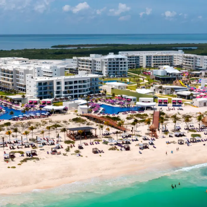 Wide aerial shot of beachfront resort with multiple pools, white buildings, and turquoise sea in the background.