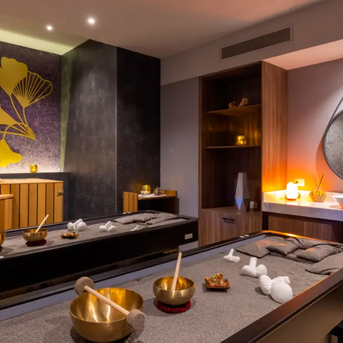 Cozy spa treatment room with massage tables, golden decor, warm lighting, and a nature-inspired wall design.