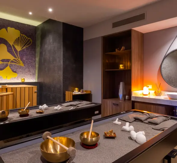Cozy spa treatment room with massage tables, golden decor, warm lighting, and a nature-inspired wall design.