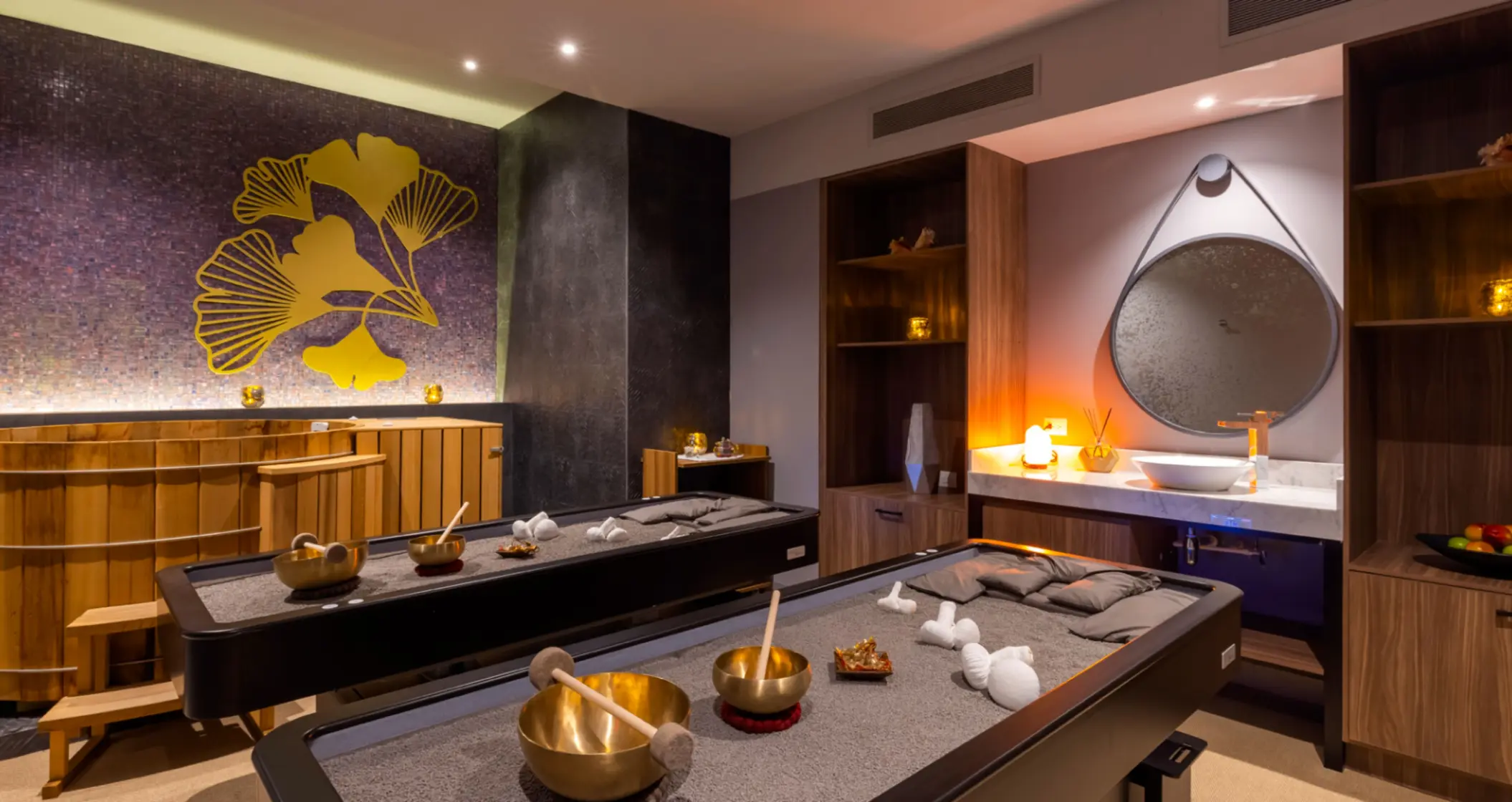 Cozy spa treatment room with massage tables, golden decor, warm lighting, and a nature-inspired wall design.