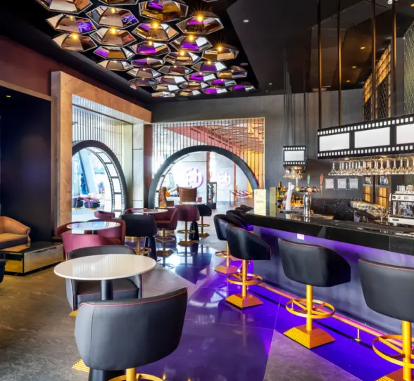 Chic bar with film-themed decor, backlit shelves, modern lounge seating, and vibrant purple and gold accents.