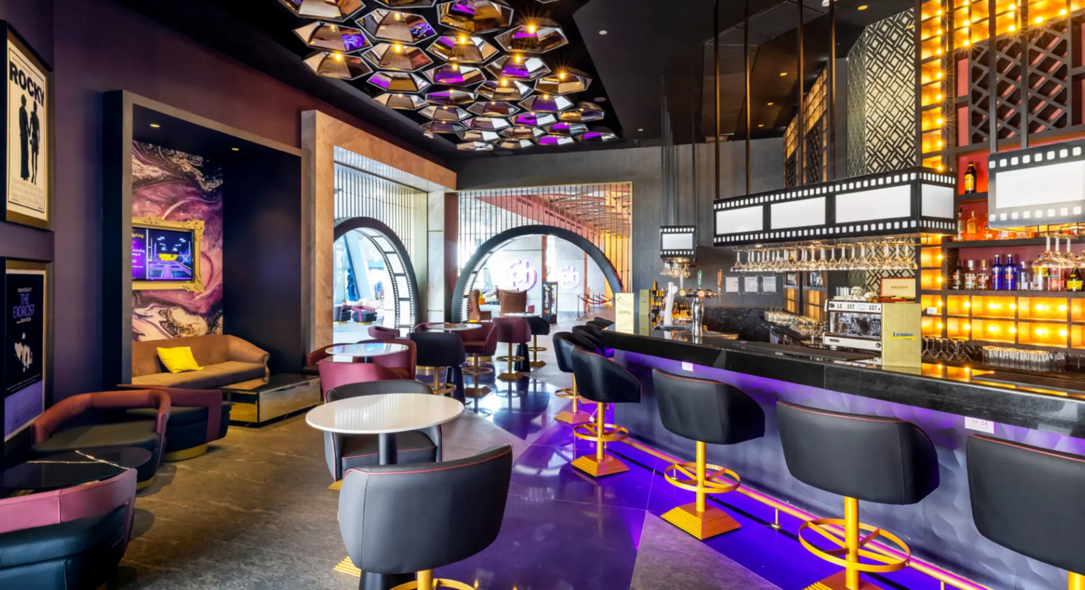 Chic bar with film-themed decor, backlit shelves, modern lounge seating, and vibrant purple and gold accents.