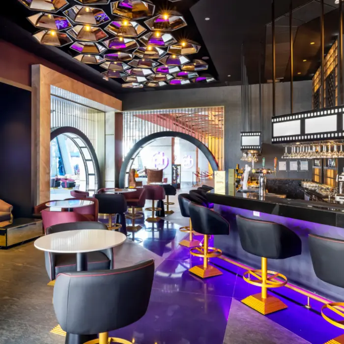 Chic bar with film-themed decor, backlit shelves, modern lounge seating, and vibrant purple and gold accents.
