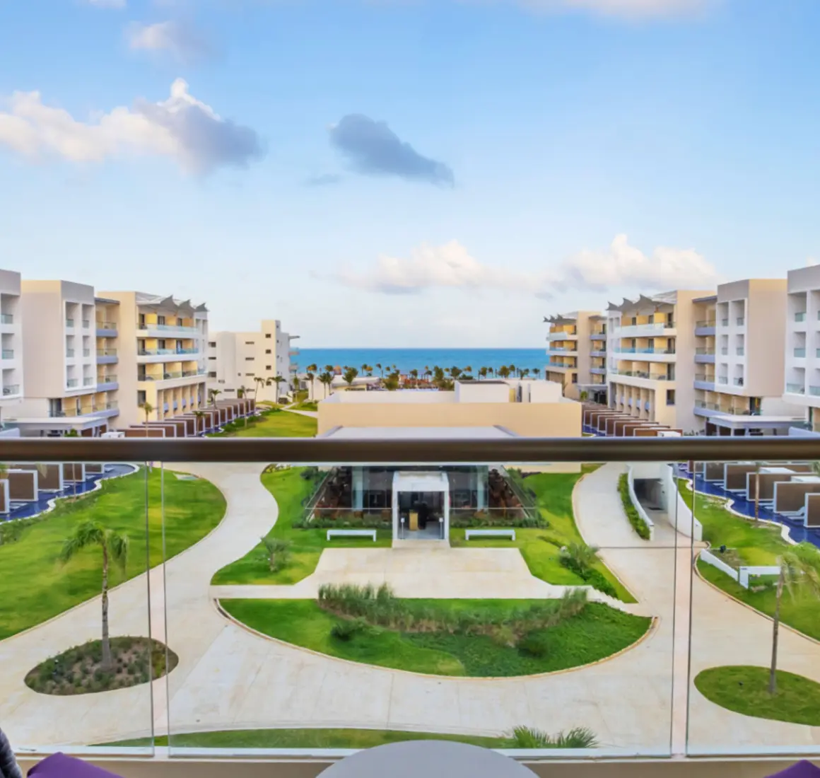 Balcony view of lush garden pathways leading to the ocean, surrounded by modern resort buildings and swim-up suites.