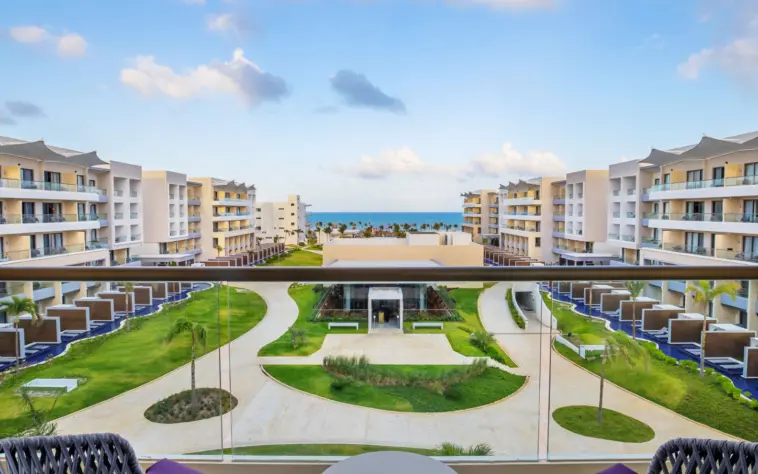 Balcony view of lush garden pathways leading to the ocean, surrounded by modern resort buildings and swim-up suites.