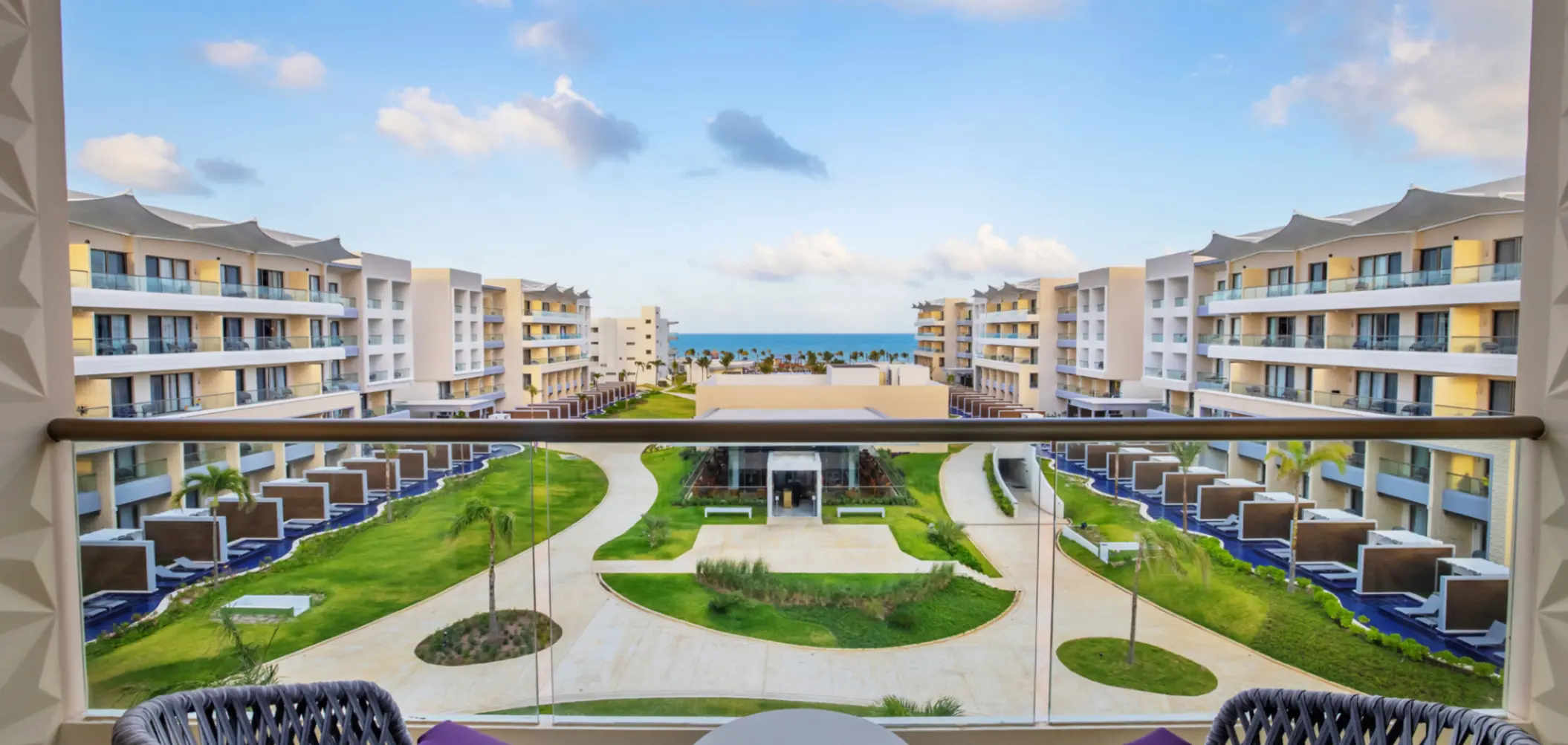 Balcony view of lush garden pathways leading to the ocean, surrounded by modern resort buildings and swim-up suites.