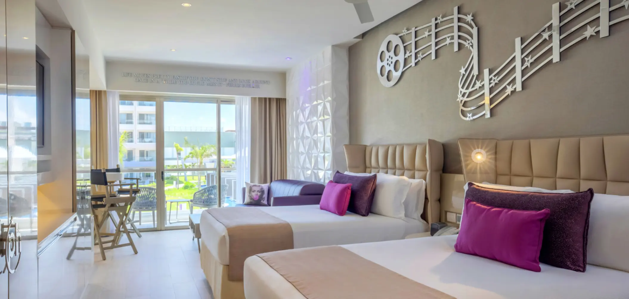 Modern resort room with double beds, balcony view, music-themed decor, and touches of purple and fuchsia pillows.
