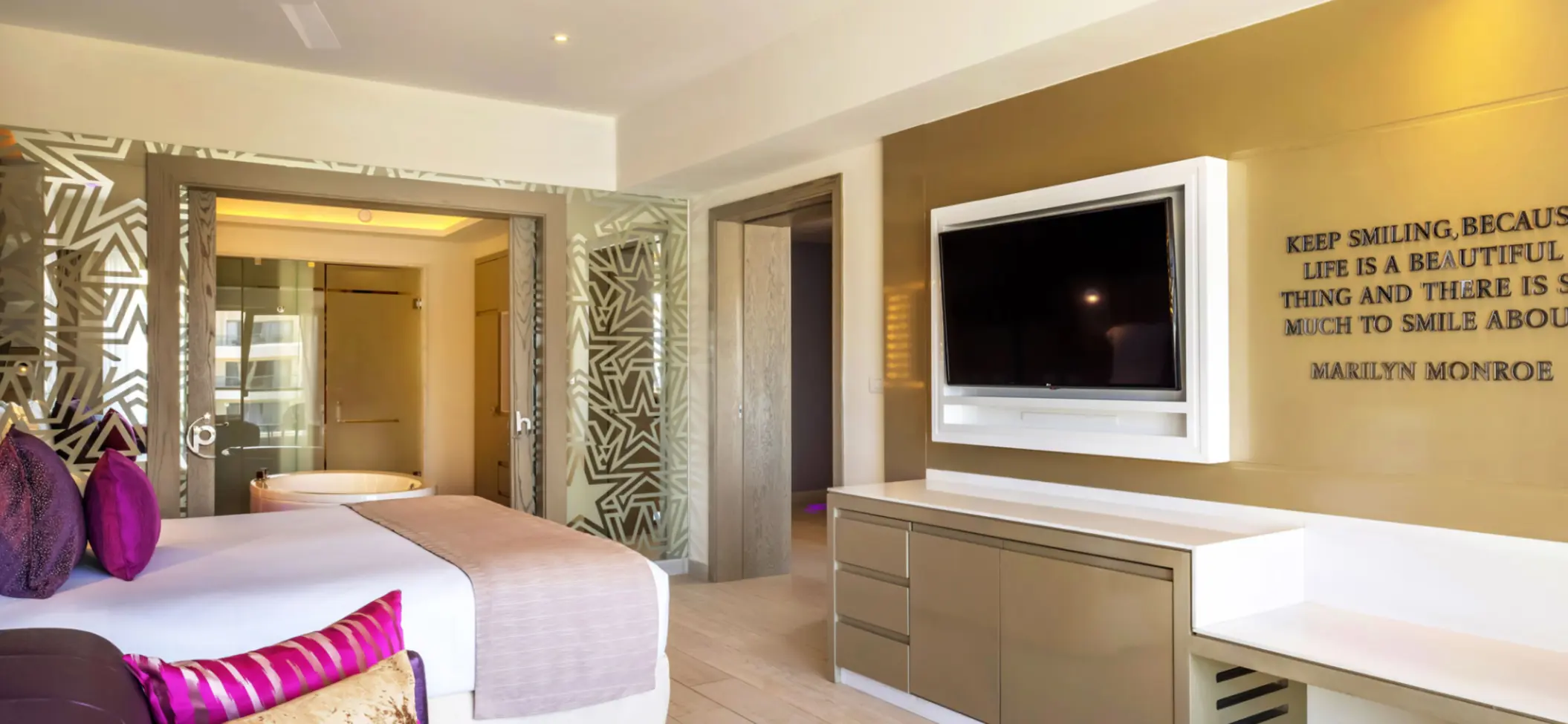 Spacious resort suite with gold accent wall, large TV, and freestanding soaking tub visible through glass panels.