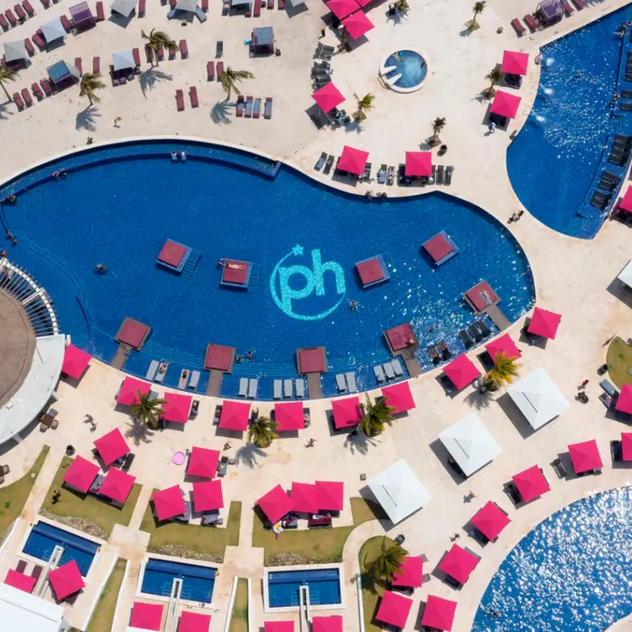 Aerial view of resort pools, pink umbrellas, beach loungers, and cabanas arranged around a central lagoon-style pool.