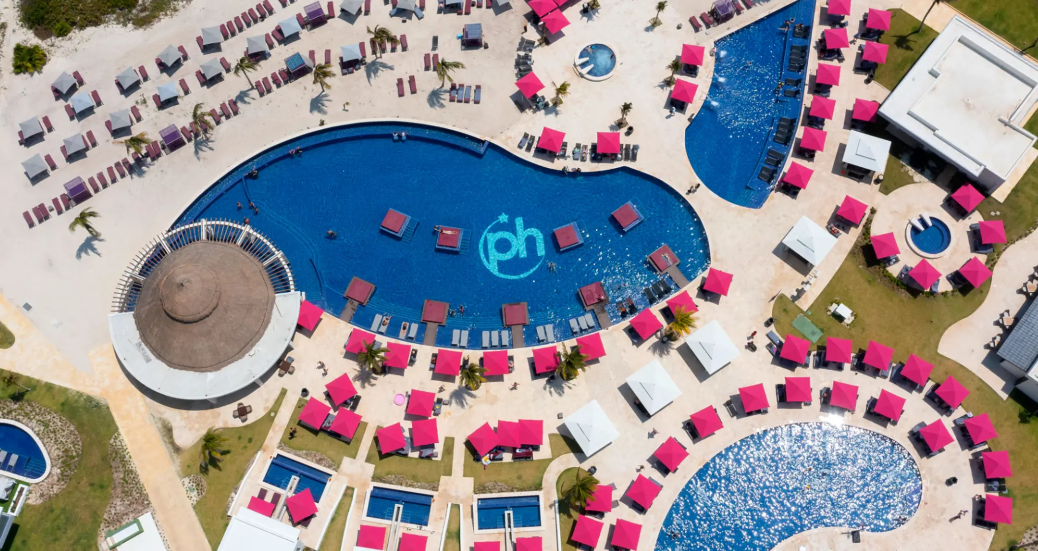Aerial view of resort pools, pink umbrellas, beach loungers, and cabanas arranged around a central lagoon-style pool.