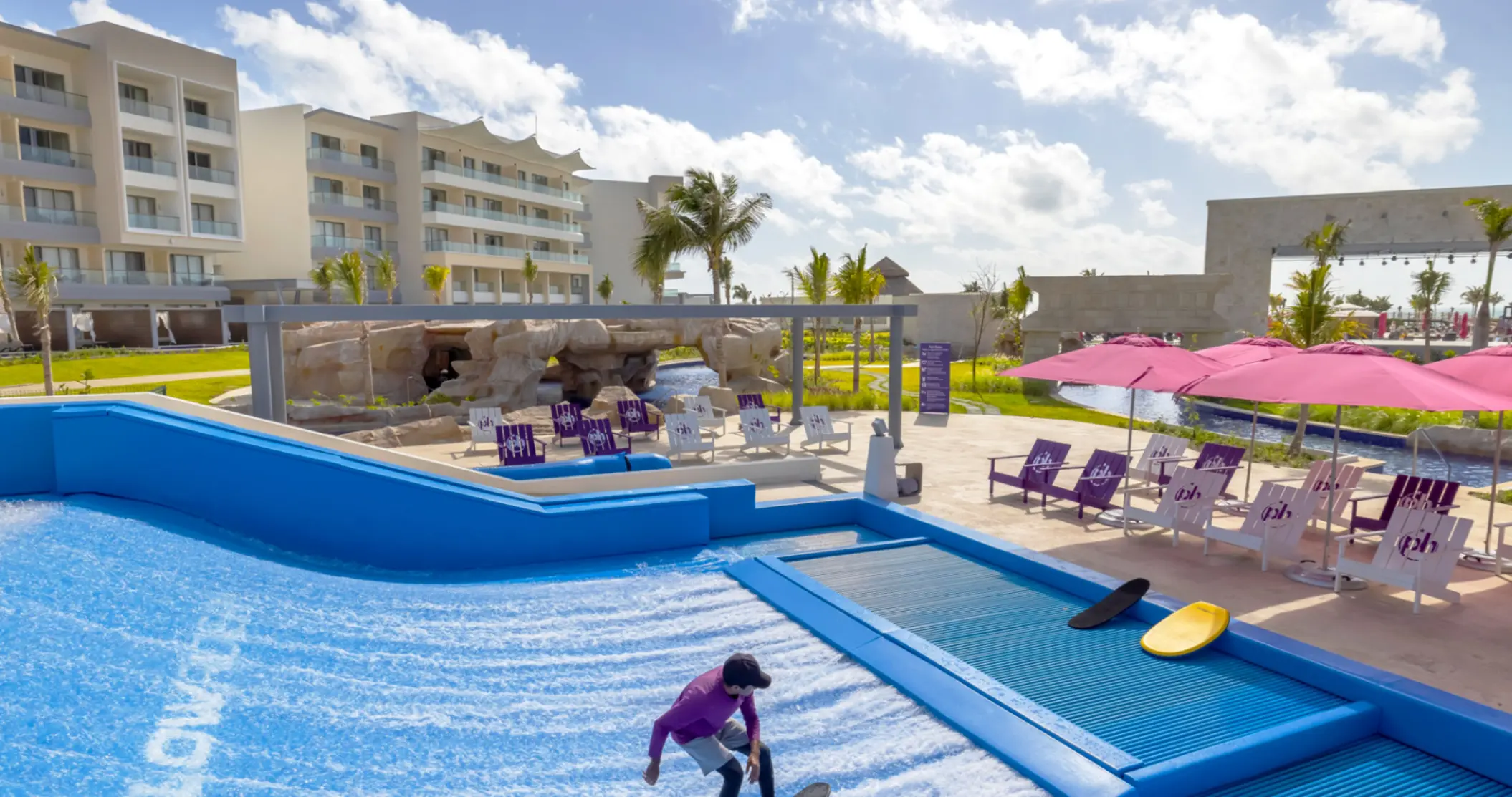 Surf simulator near poolside loungers and pink umbrellas at a tropical all-inclusive beach resort.