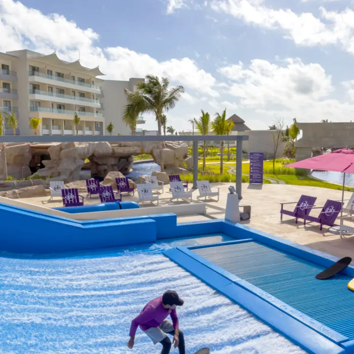 Surf simulator near poolside loungers and pink umbrellas at a tropical all-inclusive beach resort.