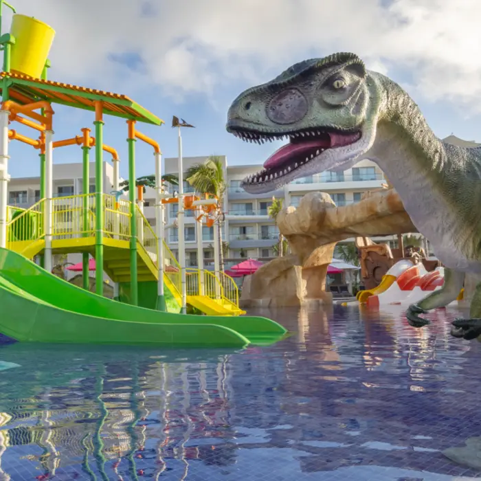 Colorful splash zone with green slides, water buckets, and dinosaur figure at a family-friendly resort water park.
