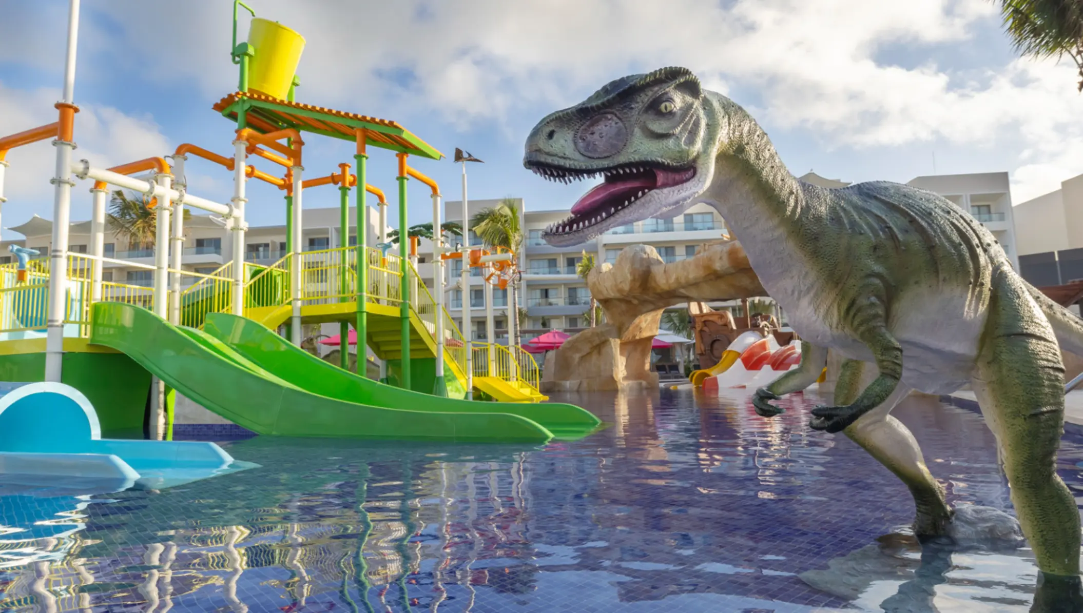 Colorful splash zone with green slides, water buckets, and dinosaur figure at a family-friendly resort water park.
