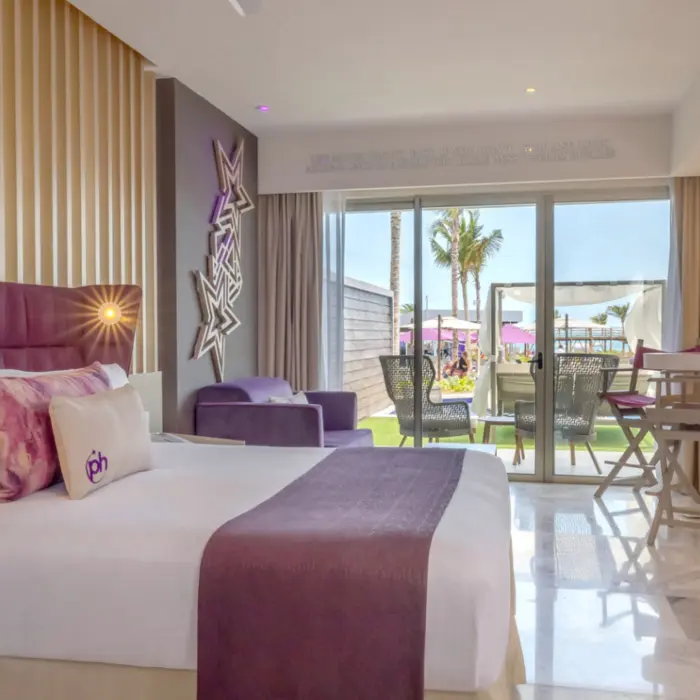 Ground-level resort room with purple accents, king bed, and furnished patio opening to lush garden and pool area.