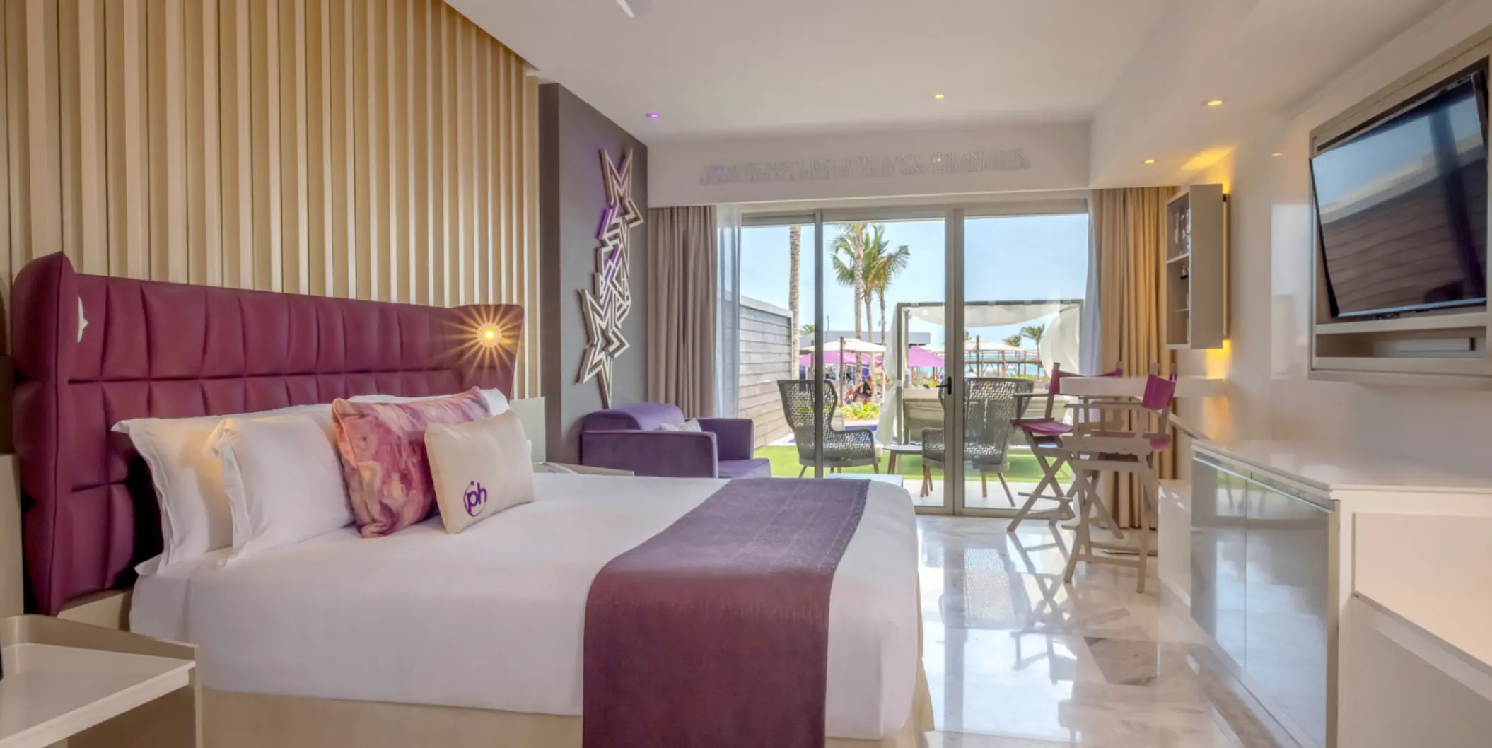 Ground-level resort room with purple accents, king bed, and furnished patio opening to lush garden and pool area.