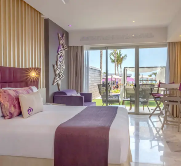 Ground-level resort room with purple accents, king bed, and furnished patio opening to lush garden and pool area.