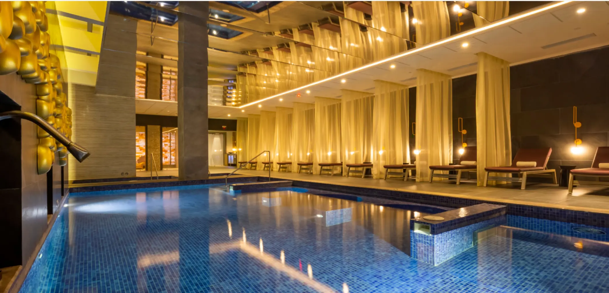 Indoor hydrotherapy pool surrounded by soft lighting, sheer curtains, and lounge chairs in an elegant spa setting.