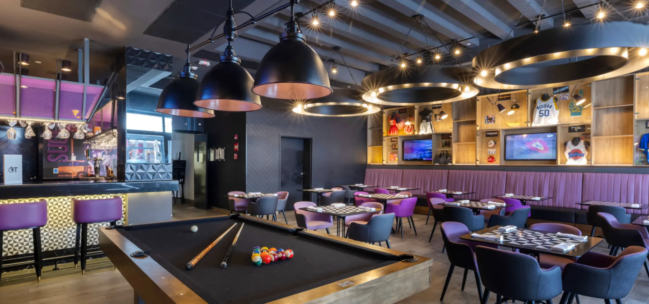 Trendy sports bar with modern decor, game tables, large TV screens, and purple-lit bar counter seating.