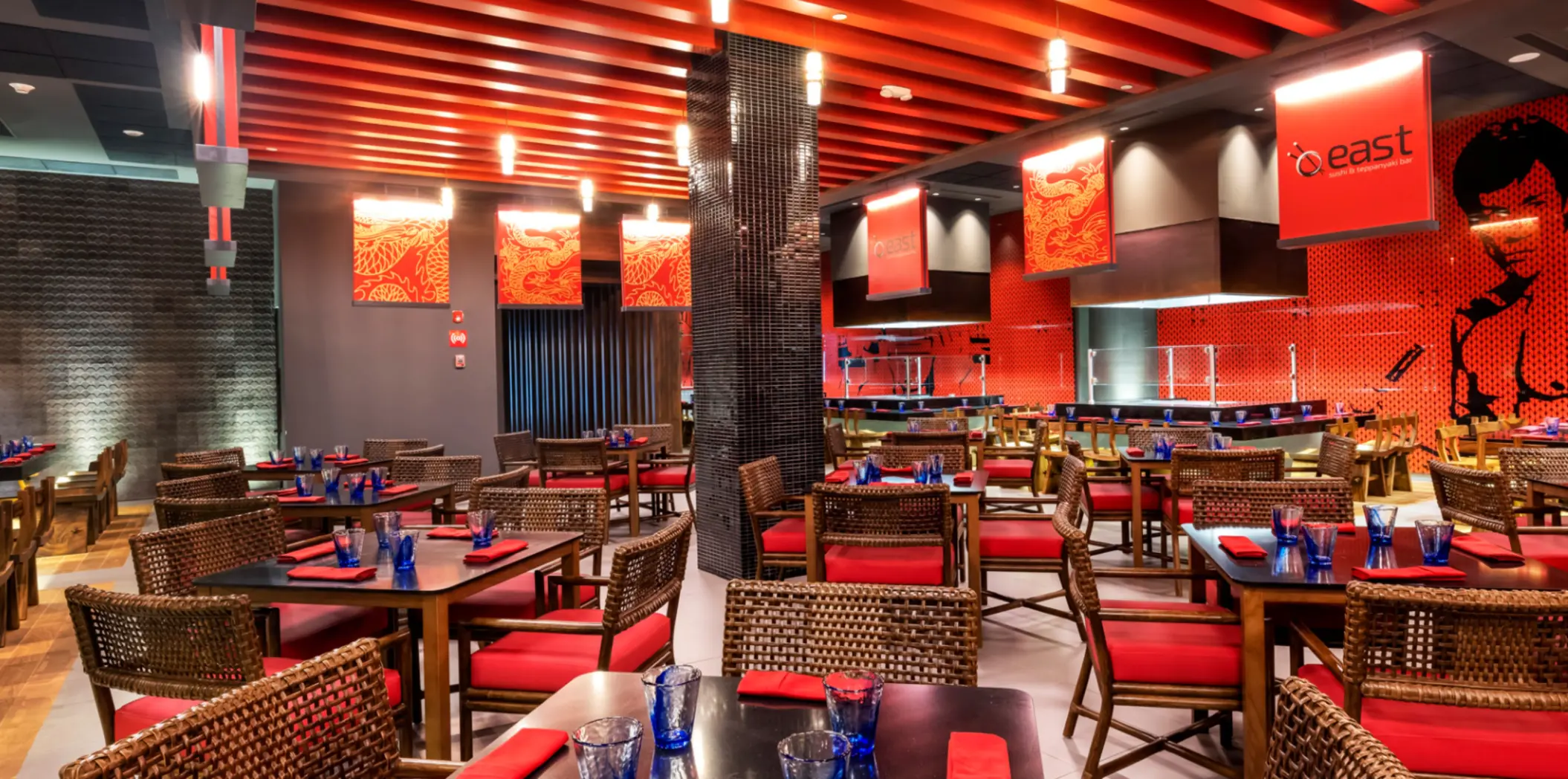 Asian restaurant with red decor, woven chairs, modern teppanyaki setup, and dragon-themed artwork on the walls.