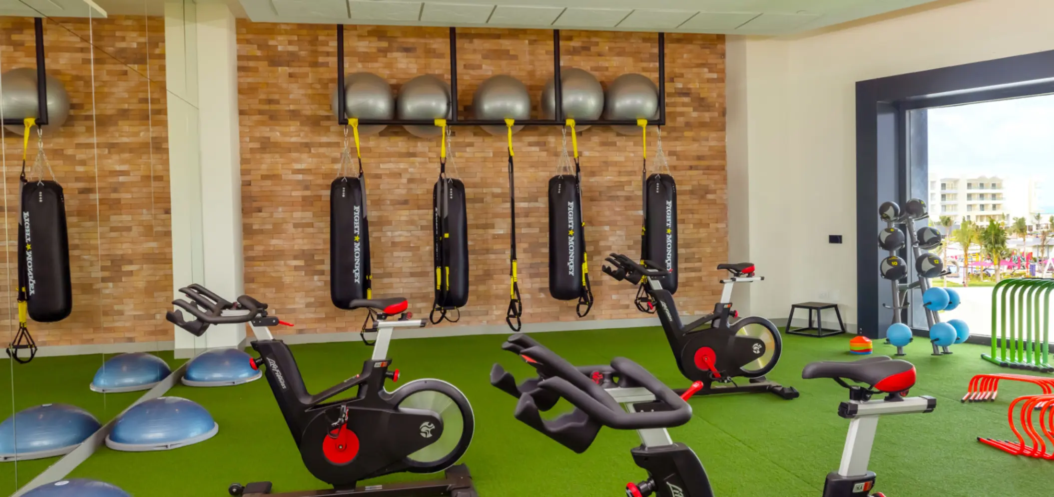 Fitness center with bikes, punching bags, and TRX equipment in a bright room with outdoor views.