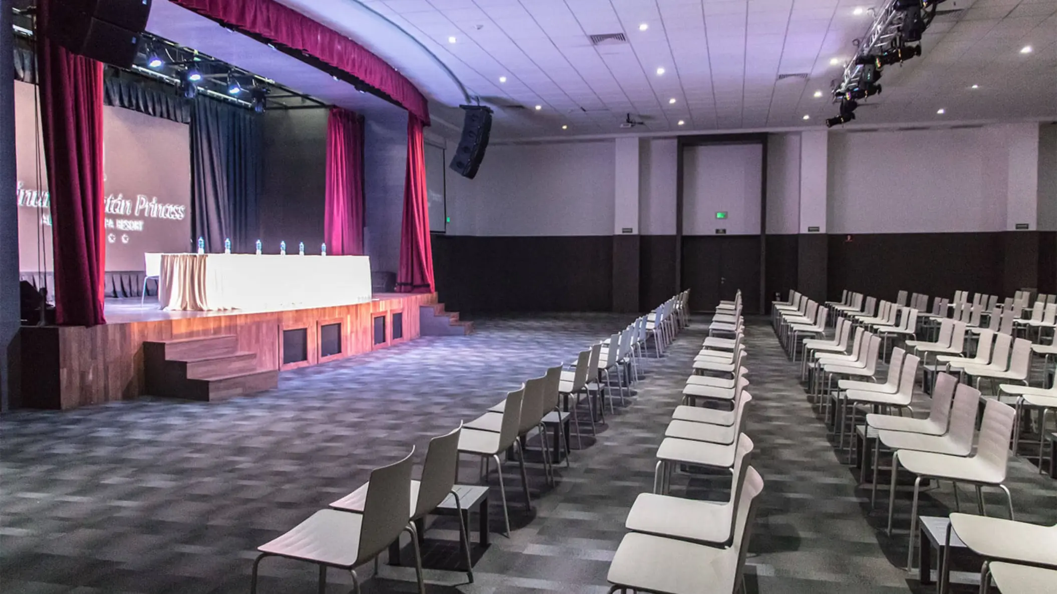 An empty conference room with rows of chairs facing a stage, featuring a head table and large screen.
