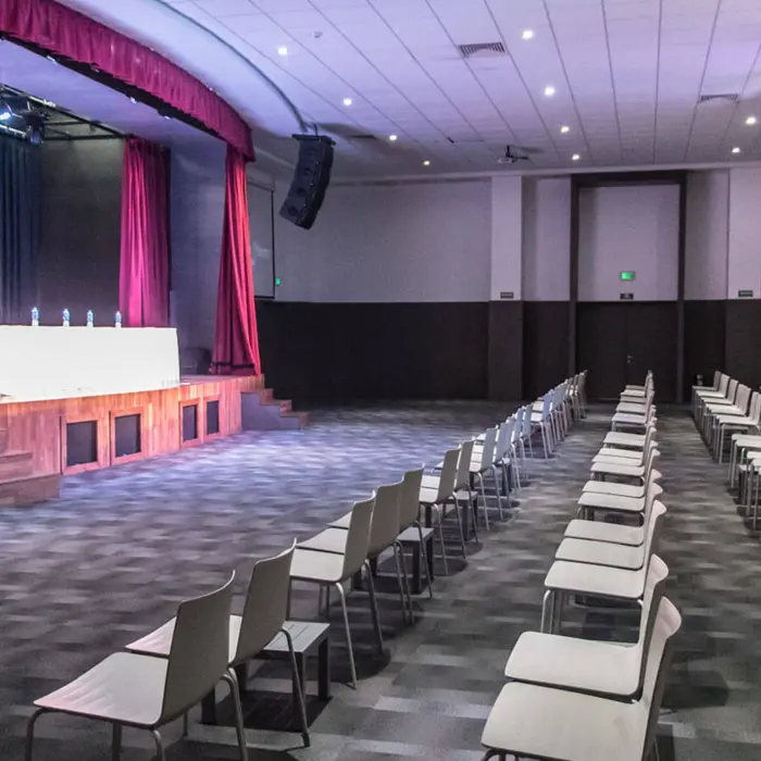 An empty conference room with rows of chairs facing a stage, featuring a head table and large screen.