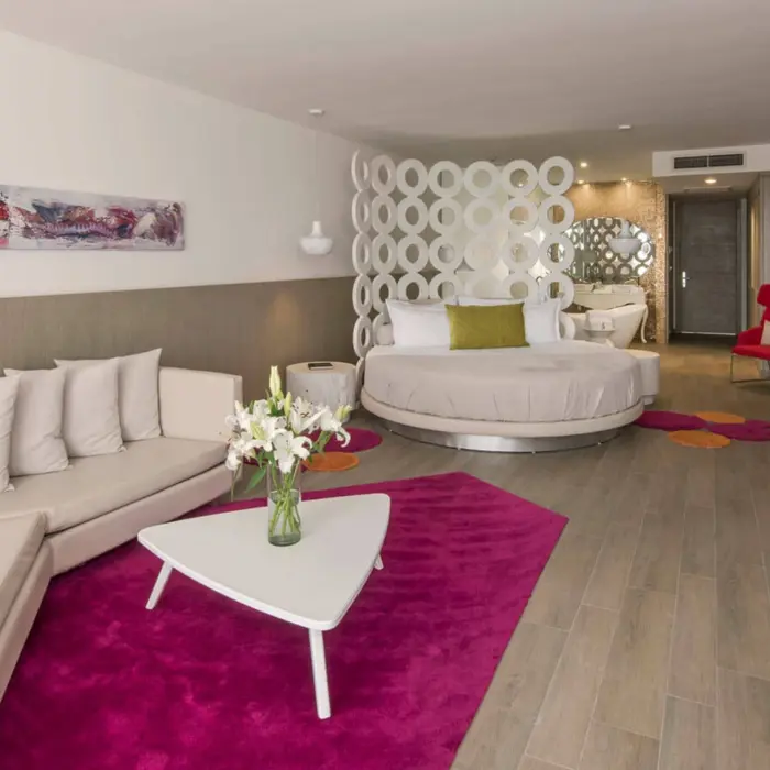 A modern Honeymoon Suite with a large circular bed, pink rug, white sofa, and a colorful abstract painting.