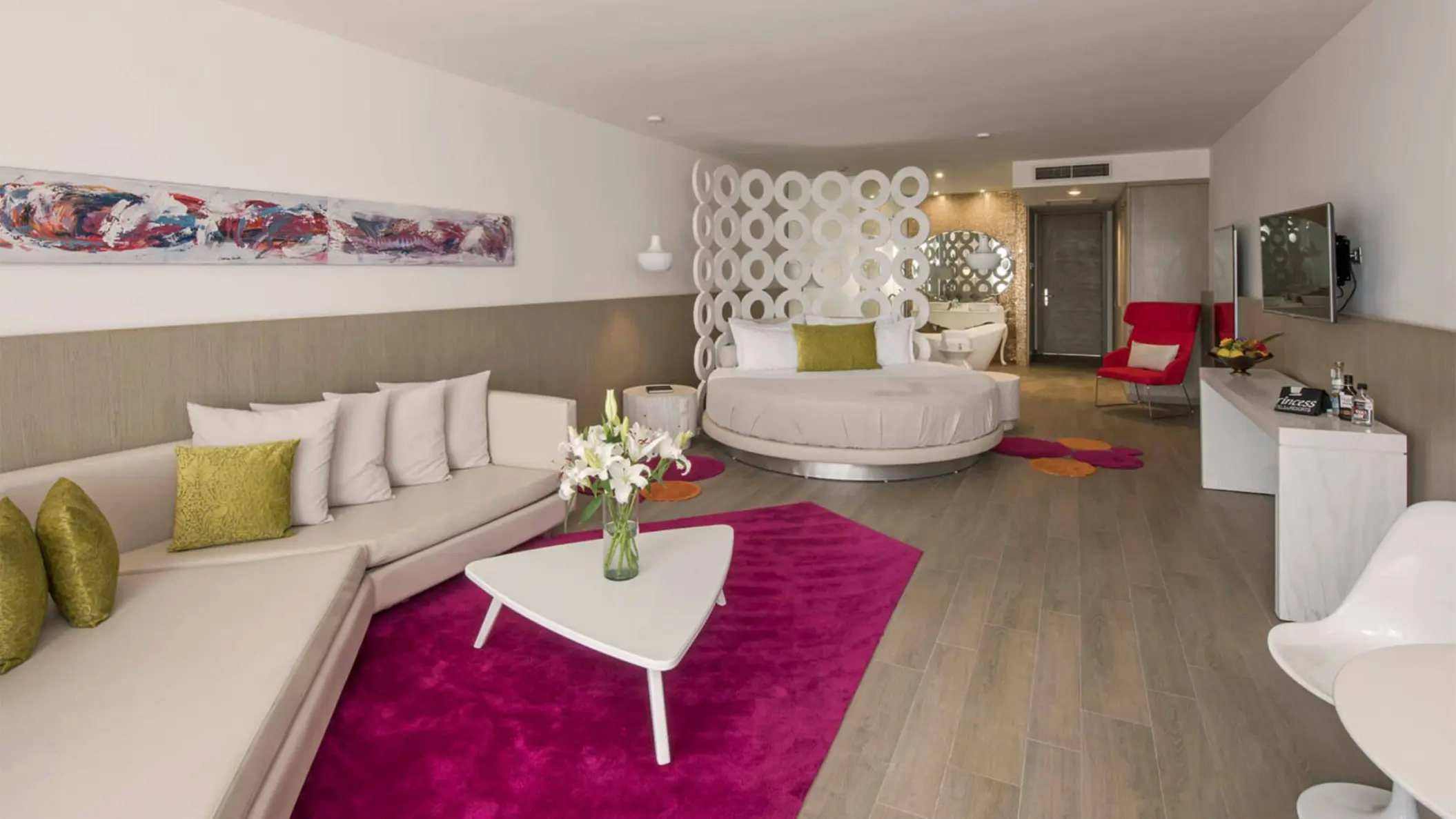 A modern Honeymoon Suite with a large circular bed, pink rug, white sofa, and a colorful abstract painting.