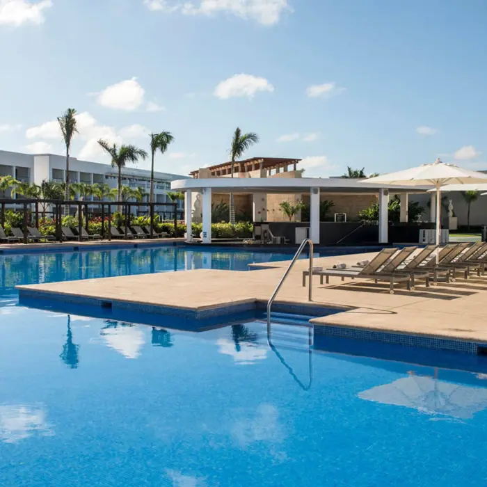 A large outdoor swimming pool area with numerous lounge chairs, palm trees, and a swim-up bar pavilion.