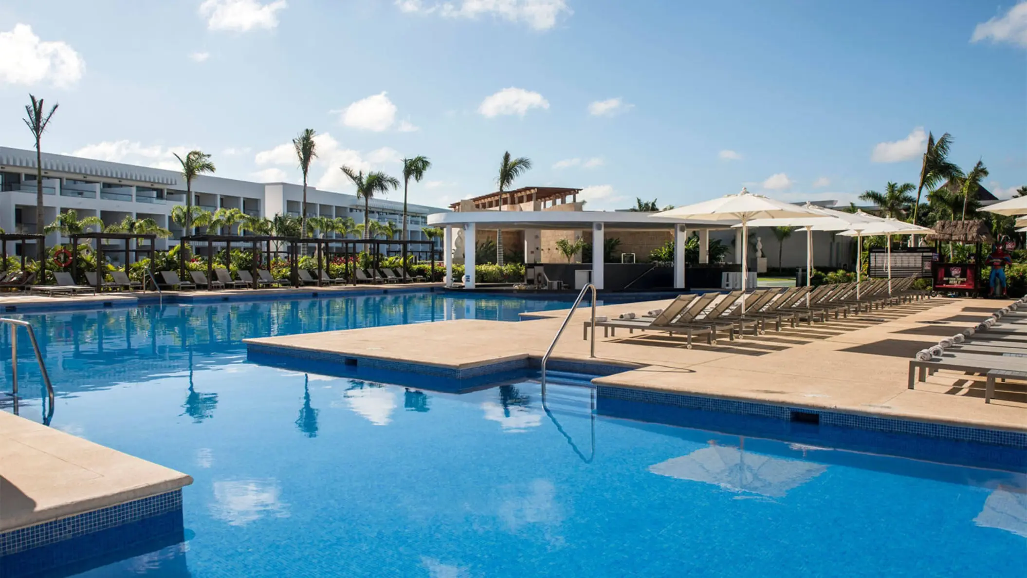 A large outdoor swimming pool area with numerous lounge chairs, palm trees, and a swim-up bar pavilion.