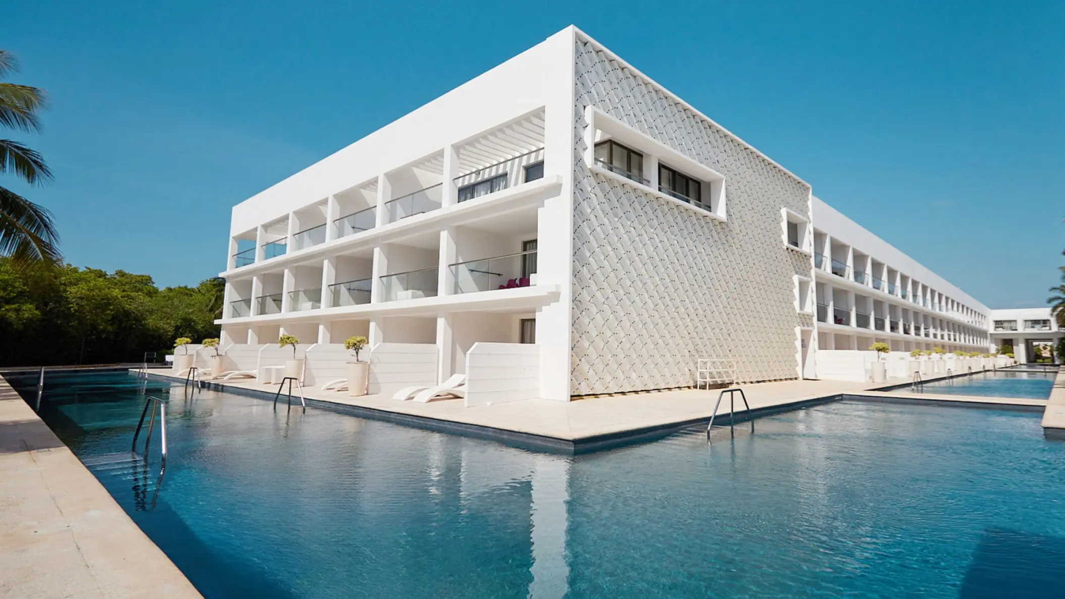 Exterior view of the hotel building overlooking a large pool, with contemporary white architecture and glass balconies.